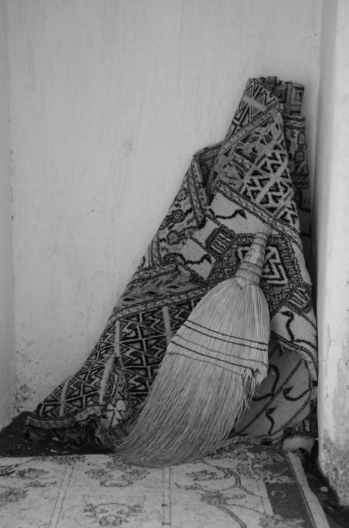 A black and white still life of a broom and rug in a simple home corner.